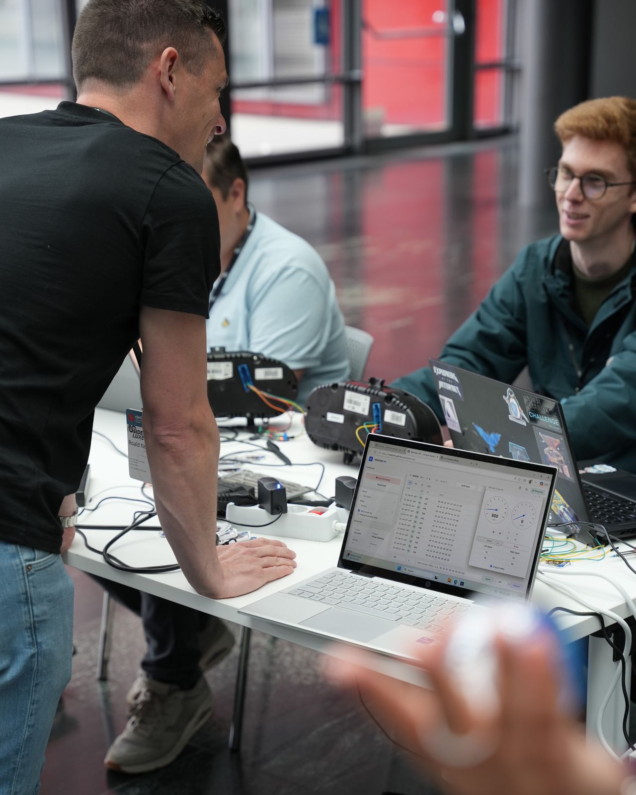 Attendees at the Car Hacking Village booth at BSides Luxembourg 2026, with instrument clusters and laptops on a workshop table.
